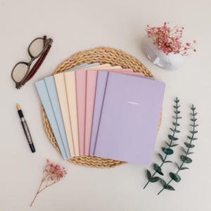 May include: A collection of pastel-colored notebooks arranged on a woven placemat. The notebooks are in shades of blue, yellow, pink, and purple. A pair of glasses, a pen, and decorative flowers are also present, creating a serene, organized aesthetic.
