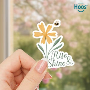 May include: A floral sticker featuring a yellow daisy with a small bee and the words "Rise & Shine." The sticker has a white background and green leaves. The Happy Hoos logo is in the top right corner.
