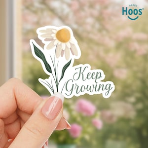 Può includere: Un adesivo a forma di margherita bianca con centro giallo e foglie verdi. L'adesivo riporta la scritta "Keep Growing" in un carattere corsivo. L'adesivo è tenuto in mano.
