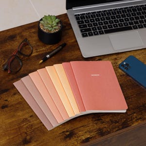 May include: A collection of notebooks in a gradient of colors, from light brown to coral, fanned out on a wooden desk. The word "NOTEBOOK" is printed on the cover of one notebook. A laptop, phone, glasses, pen, and a small potted plant are also on the desk.