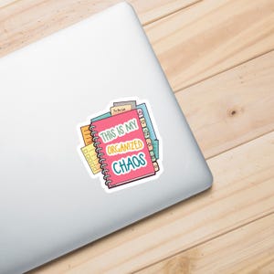 A laptop with a colorful vinyl sticker featuring an illustration of stacked spiral notebooks and planners with tabs. The main text reads 'THIS IS MY ORGANIZED CHAOS' in white, yellow, and teal lettering on a pink notebook cover. The sticker shows various planning elements like to-do lists, checkboxes, and organizational tabs in bright colors including pink, yellow, green, and blue.
