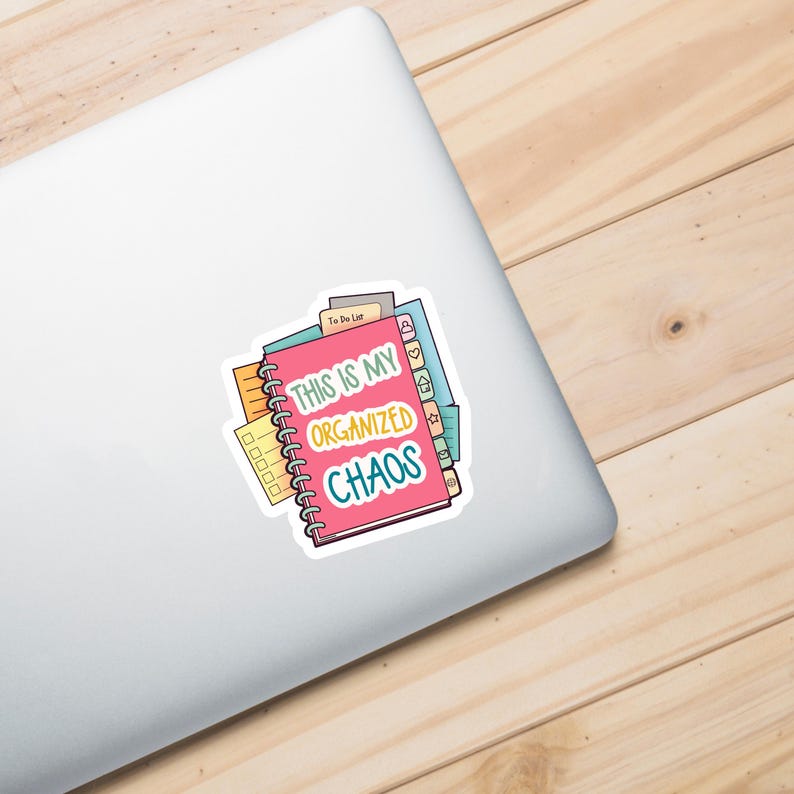 A laptop with a colorful vinyl sticker featuring an illustration of stacked spiral notebooks and planners with tabs. The main text reads 'THIS IS MY ORGANIZED CHAOS' in white, yellow, and teal lettering on a pink notebook cover. The sticker shows various planning elements like to-do lists, checkboxes, and organizational tabs in bright colors including pink, yellow, green, and blue.