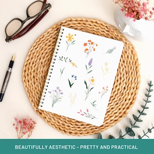 May include: A white spiral-bound notebook with floral illustrations, including daisies, lavender, and other wildflowers. The notebook is on a woven placemat, with a pen and glasses nearby. The text at the bottom reads: BEAUTIFULLY AESTHETIC - PRETTY AND PRACTICAL.