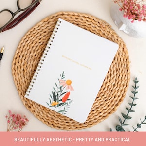 May include: A white spiral-bound notebook with floral illustrations and the text "Write your journey, cultivate joy." The notebook is on a woven placemat, with glasses and a pen nearby. The bottom of the image reads "BEAUTIFULLY AESTHETIC - PRETTY AND PRACTICAL."