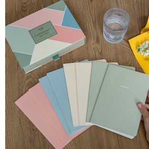 May include: A notebook gift set with a geometric design box in pink, teal, and green. The set includes several notebooks in pastel colors: pink, blue, cream, and green. A glass of water and a yellow flower arrangement are also visible.