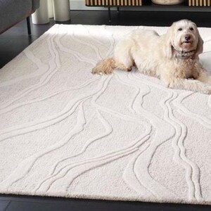 May include: A cream-coloured rectangular rug with a raised, flowing pattern. A light-coloured dog is lying on the rug. The rug's design features a series of curved lines, creating a textured surface. The rug is placed on a dark floor.