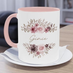 May include: White ceramic mug with a pink interior and handle, featuring floral illustrations in shades of pink and brown. The name "Ginie" is written in cursive script. The mug sits on a white saucer.