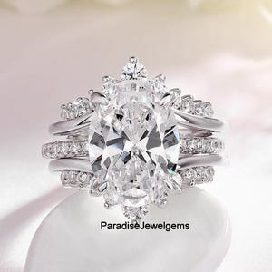Peut inclure: Un ensemble de trois bagues en argent avec une grosse pierre précieuse claire de taille ovale au centre. La bague est entourée de petites pierres précieuses claires. Le texte "ParadiseJewelgems" est visible.