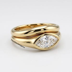 Könnte beinhalten: Ein goldfarbenes Ring-Set. Der Verlobungsring hat einen Diamanten im Marquise-Schliff. Das Band ist glatt und abgerundet. Der Ring ist vor einem weißen Hintergrund platziert. Der Text "ParadiseJewelgems" ist sichtbar.