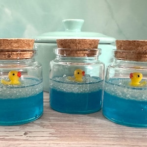 May include: Three small glass jars with cork stoppers, each containing blue liquid and a yellow rubber duck. The jars are arranged in a row, with a light blue background.