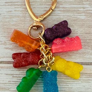 May include: A gold-colored keychain with a heart-shaped clasp, holding a cluster of colorful gummy bear charms. The charms are in various colors, including orange, red, green, blue, yellow, pink, and purple, and have a glittery finish.