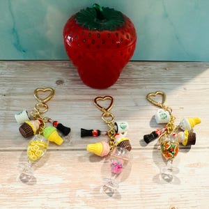 May include: A red strawberry-shaped container with a green stem sits on a wooden surface. Three keychains with gold heart-shaped clasps hang below. Each keychain has miniature food charms, including cupcakes, ice cream cones, and soda bottles.