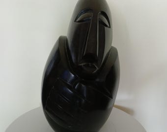 May include: A dark, abstract sculpture of a face and torso. The sculpture is a solid, glossy black, with carved features including eyes, a nose, and a mouth. The figure appears to be holding its arms across its chest.