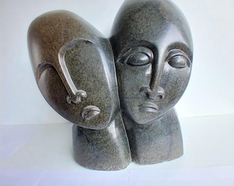 May include: A gray stone sculpture depicting two stylized human faces. One face is turned towards the other, with closed eyes and a serene expression. The sculpture has a smooth, polished finish and is set against a white background.