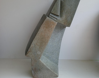 May include: A gray stone sculpture of a stylized human head and torso. The sculpture has a rectangular head with a circular eye and a triangular nose. The torso is angular with a textured surface. The sculpture is a unique piece of art.