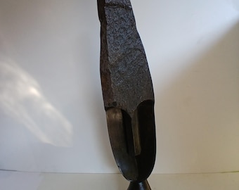May include: A dark brown, abstract sculpture with a rough, textured surface. The sculpture has a tall, elongated shape with a hollowed-out section resembling a face. It is mounted on a small, rounded base, creating a striking artistic piece.