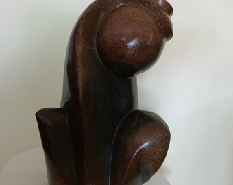 May include: A brown abstract sculpture with a smooth, polished surface. The sculpture has a rounded top and a curved, flowing design. It is placed on a small, white, round table.