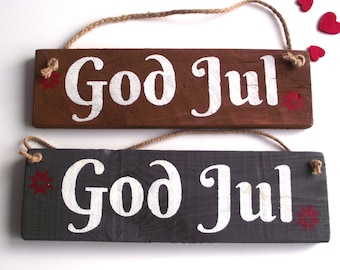 May include: Two wooden signs with the text "God Jul" in white paint. The signs are brown and gray and have red stars painted on them. The signs are hanging from a twine rope.