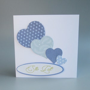 May include: A white greeting card featuring four blue heart shapes of varying sizes. The hearts have different patterns, including polka dots and a geometric design. The card also has an oval with the text "Sto Lat!"