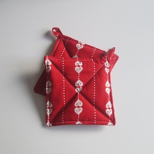 May include: Two red square pot holders with white heart and dot patterns. Each pot holder has a small loop for hanging. The quilted design adds texture to these kitchen accessories.