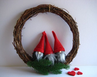 May include: A brown twig wreath with three red felt gnomes with white beards and black felt bodies. The gnomes are sitting on a bed of green pine sprigs. There are three red felt hearts on the white surface below the wreath.