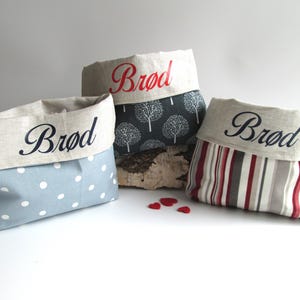 May include: Three fabric bread baskets, each with the word "Brød" embroidered on the top. The baskets feature various patterns: blue polka dots, a grey tree design, and red and grey stripes. Small red heart-shaped decorations are scattered in front.