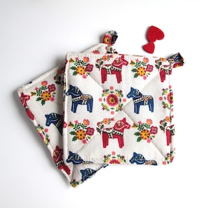 May include: Two white cotton pot holders with a red, blue and yellow floral pattern and a design of a red and blue Dala horse.