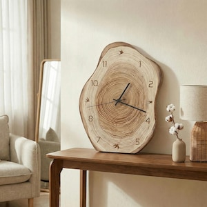 Puede incluir: Un reloj de pared de madera con una forma natural y orgánica. La esfera del reloj presenta los anillos de un tronco de árbol, con manecillas y números negros. Aves decorativas están colocadas en las posiciones de las 1, 4, 7 y 10 en punto. El reloj está montado en una pared clara.