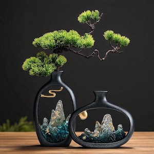 Puede incluir: Dos jarrones de cerámica negra con un recorte ovalado, que presentan paisajes de montaña en miniatura y detalles dorados. Un jarrón tiene un detalle dorado en forma de curva, el otro una esfera dorada. Ramas de pino artificiales verdes se extienden de los jarrones.