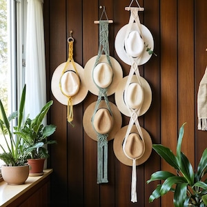 May include: Hats displayed on a wooden wall, suspended by macrame hangers in yellow, green, and white. The hats are a mix of white and tan. Potted plants are visible in the foreground, adding a touch of greenery.