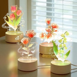 Pode incluir: Quatro lâmpadas decorativas LED em forma de flores. Cada lâmpada apresenta um design floral diferente: tulipas, rosas e lírios-do-vale. As flores são feitas de acrílico transparente e são iluminadas por uma luz quente da base.