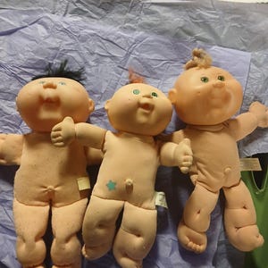 May include: Three vintage Cabbage Patch Kids dolls. The dolls have soft, peach-coloured skin, green eyes, and varying hair colours. One doll has black hair, one has red hair, and the other has blonde hair. They are lying on a purple surface.