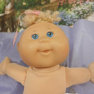 May include: A vintage Cabbage Patch Kid doll with a peach-colored skin tone, blue eyes, and blonde curly hair tied with a pink ribbon. The doll is wearing a peach-colored onesie and has a tag with the initials "CPK".