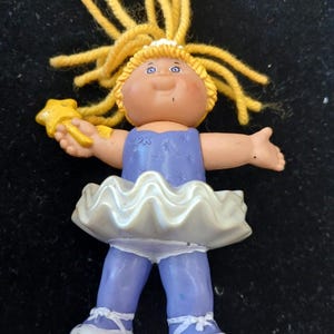 May include: A vintage Cabbage Patch Kids ballerina doll. The doll has yellow yarn hair, blue eyes, and a light skin tone. She wears a blue leotard, a white tutu, and white ballet shoes. The doll holds a yellow star-shaped wand.