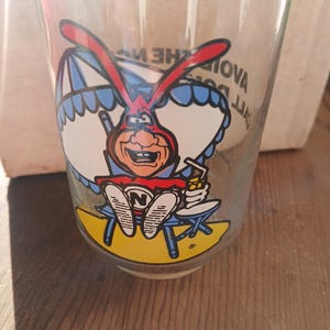 May include: Clear glass with a cartoon character design. The character has a red head, blue ears, and is seated at a small table. The glass has a yellow base and a blue and white awning design.