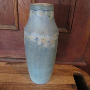 May include: A tall, cylindrical ceramic vase with a matte, mottled light blue and grey finish. The vase features a hand-painted floral pattern in soft white and pale yellow tones encircling the upper section and the mid-body area.