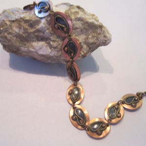 Op de afbeelding: Een koperen en zilveren armband met een uniek ontwerp met een reeks ovale schakels met ingewikkelde patronen.