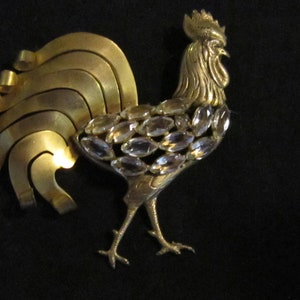 Op de afbeelding: Een gouden haan broche met een gedetailleerd ontwerp en heldere strass steentjes. De staartveren van de haan zijn gemaakt van meerdere gebogen metalen stukken.