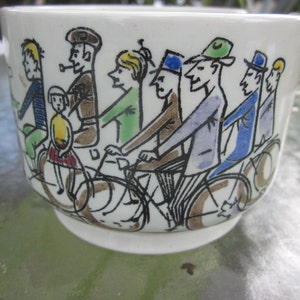 Może przedstawiać: Biała ceramiczna filiżanka do herbaty z fantazyjną ilustracją ludzi na rowerach. Postacie przedstawione są w różnych kolorach, w tym niebieskim, zielonym i brązowym, z czarnymi konturami. Filiżanka ma zaokrąglony kształt i mały uchwyt.