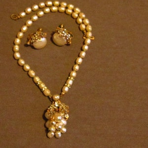 May include: A vintage jewelry set featuring a pearl necklace with a cluster pendant, and matching pearl earrings. The necklace and earrings have gold-toned accents and are set against a brown background.