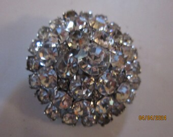 May include: A round, silver-toned brooch densely covered with clear, faceted rhinestones. The sparkling stones are arranged in a clustered pattern, creating a dazzling, eye-catching accessory. The brooch is likely vintage or costume jewelry.