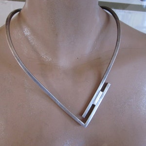 May include: A silver necklace with a geometric design. The necklace is made of a single piece of metal that is bent into a V-shape. The ends of the necklace are connected by a small bar.