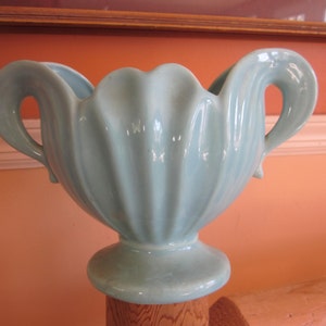 May include: A light blue ceramic vase with two handles and a fluted design. The vase is sitting on a wooden surface.