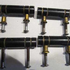 Puede incluir: Juego de cuatro tiradores de armario negros vintage con detalles de latón. Cada tirador presenta un mango cilíndrico negro con anillos de latón y un tornillo roscado. Ideal para proyectos de restauración de muebles.