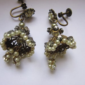 Puede incluir: Un par de pendientes de tono dorado con un racimo de perlas blancas y diamantes de imitación transparentes. Los pendientes tienen un cierre de clip.