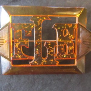 May include: A vintage amber-colored plastic brooch with gold glitter embedded in the letters "E.B."