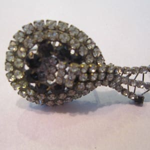 Puede incluir: Un broche de plata y negro con una pieza central grande de forma ovalada cubierta de diamantes de imitación. La pieza central está rodeada por un diseño más pequeño e intrincado de diamantes de imitación y metal.