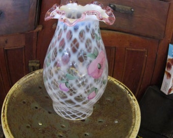 Pode incluir: Um vaso de vidro branco vintage com um design de treliça e detalhes florais rosa. O vaso tem uma borda com babados com uma borda rosa. O vaso está sobre uma superfície metálica.
