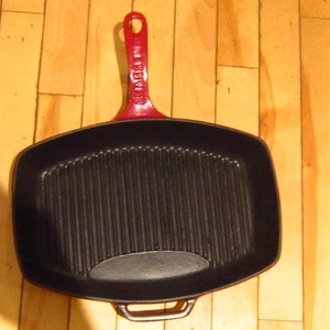 May include: A black cast iron grill pan with a red handle. The pan has a rectangular shape and a ridged cooking surface. The handle is red, and the pan is on a wooden surface.