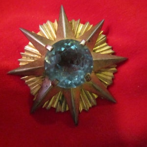 May include: A gold starburst brooch with a large, faceted, light blue gemstone in the center.